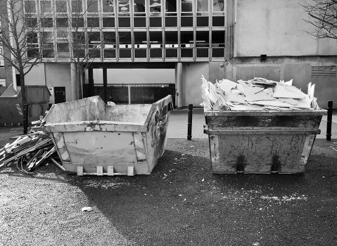 The image shows two large industrial waste skips positioned on a paved street or driveway in an urban area. The skip on the left appears to contain broken concrete and rubble with a rough, uneven surface and grey coloration, indicating construction or demolition debris. The skip on the right is filled with crumpled and torn white and beige papers or cardboard, stacked haphazardly and spilling over the edges, suggesting paper waste or packaging material. Both skips are situated in front of a modern building facade with a concrete or metal exterior, large glass windows, and a gated entrance visible in the background. The surrounding environment includes a few leafless trees and small bushes on the left side, with some dirt and small debris scattered on the ground nearby. The lighting appears natural but overcast, casting soft shadows and a muted tone across the scene, which aligns with typical rubbish removal operations in Islington or nearby areas. The image is associated with Business Waste Removal Islington, specializing in rubbish collection and disposal services.