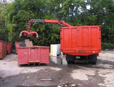 The image shows an industrial waste collection scene outdoors, where a red garbage truck equipped with a mechanical arm and hydraulic grab attachment is in the process of loading waste into a large, open-top red skip positioned on an asphalt surface. The waste being lifted appears to include various materials, possibly including cardboard, plastics, or mixed rubbish, which are partially visible at the top of the skip. The surrounding environment features a background of lush green trees and foliage, indicating a location near a wooded or green area. The scene appears to take place during daylight hours with natural light illuminating the scene evenly. Visible on the ground are scattered bits of litter, debris, and small stones. Business Waste Removal Islington handles rubbish collection and waste disposal services, and this setup is representative of their typical operations for managing commercial and industrial waste in urban environments near Islington and the surrounding postcode areas.