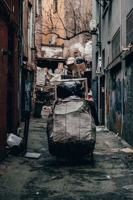 A narrow urban alleyway cluttered with accumulated waste materials, featuring a large, worn black trash bin with visible creases and patches, positioned centrally on a dark, uneven pavement. Behind th