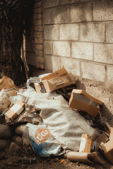 A narrow urban alleyway cluttered with accumulated waste materials, featuring a large, worn black trash bin with visible creases and patches, positioned centrally on a dark, uneven pavement. Behind th