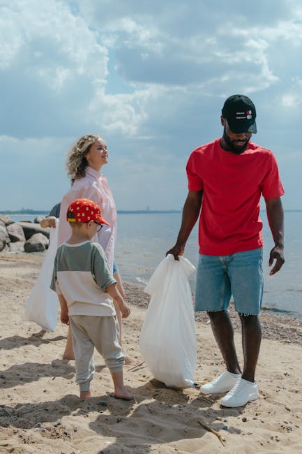 A group of three people cleaning up a sandy beach area near a body of water. A man dressed in a red t-shirt, light blue denim shorts, white sneakers, and a black cap is holding a large white plastic b