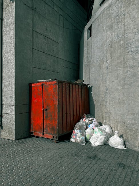 A commercial waste collection vehicle, predominantly white with visible rust and wear on its rear compartment, is positioned on a narrow cobblestone street beside a row of aged, multi-storey buildings